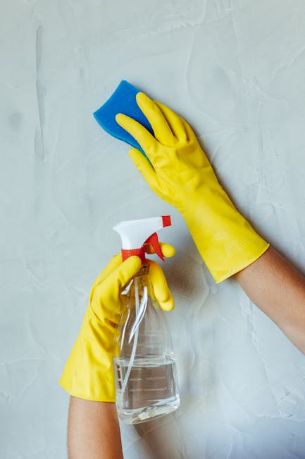 A person wearing bright yellow rubber gloves is cleaning a white wall with a blue sponge in one hand while holding a spray bottle filled with clear cleaning solution in the other hand. The scene is set indoors, with the person actively engaged in house cleaning tasks as part of a home relocation process. The background features a plain, textured white wall, and the lighting is neutral and even, highlighting the cleaning activity. This image reflects the preparation and packing stages involved in furniture transport and moving services, emphasizing thorough cleaning and organisation typical of professional removals, such as those offered by Man with Van Wormwood Scrubs during house removals and moving logistics.