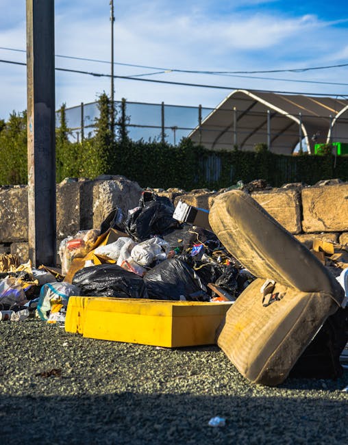 A pile of household waste and discarded items located on a gravel surface outside, including black and yellow plastic rubbish bags, an old beige upholstered armchair with visible wear and dirt, and a large yellow plastic container. Some waste is spilling out of the bags and the container, with crushed cardboard boxes and packaging materials mixed in. In the background, a stone wall and green bushes are visible, along with utility poles and overhead power lines under a partly cloudy sky. The scene is outdoors, near a fencing area, and may be associated with bulk waste disposal as part of a home relocation or furniture transport process, reflecting typical steps in house removals managed by companies like Man with Van Wormwood Scrubs.
