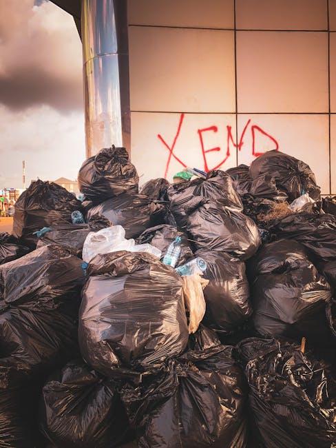 A large pile of black plastic garbage bags, filled with household waste, positioned outdoors against a tiled wall with red graffiti spelling 'XEN'. Some bags contain visible plastic bottles and packaging materials, indicating mixed waste. The bags are stacked on pavement, suggesting they are ready for disposal or collection. The scene is illuminated by natural light, likely in the late afternoon, with a cloudy sky visible in the upper left portion of the image. This arrangement of waste could be part of a home relocation or clearance process handled by Man with Van Wormwood Scrubs, illustrating the type of bulky waste disposal service they provide for clients near Wormwood Scrubs area.