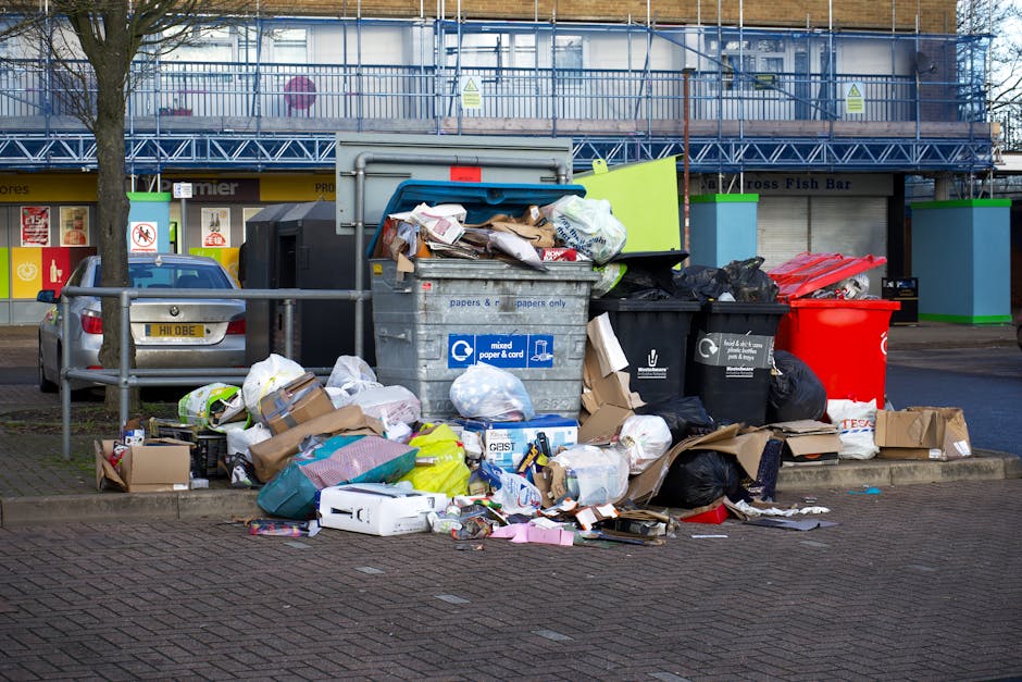 A pile of household waste and discarded items located on a gravel surface outside, including black and yellow plastic rubbish bags, an old beige upholstered armchair with visible wear and dirt, and a large yellow plastic container. Some waste is spilling out of the bags and the container, with crushed cardboard boxes and packaging materials mixed in. In the background, a stone wall and green bushes are visible, along with utility poles and overhead power lines under a partly cloudy sky. The scene is outdoors, near a fencing area, and may be associated with bulk waste disposal as part of a home relocation or furniture transport process, reflecting typical steps in house removals managed by companies like Man with Van Wormwood Scrubs.