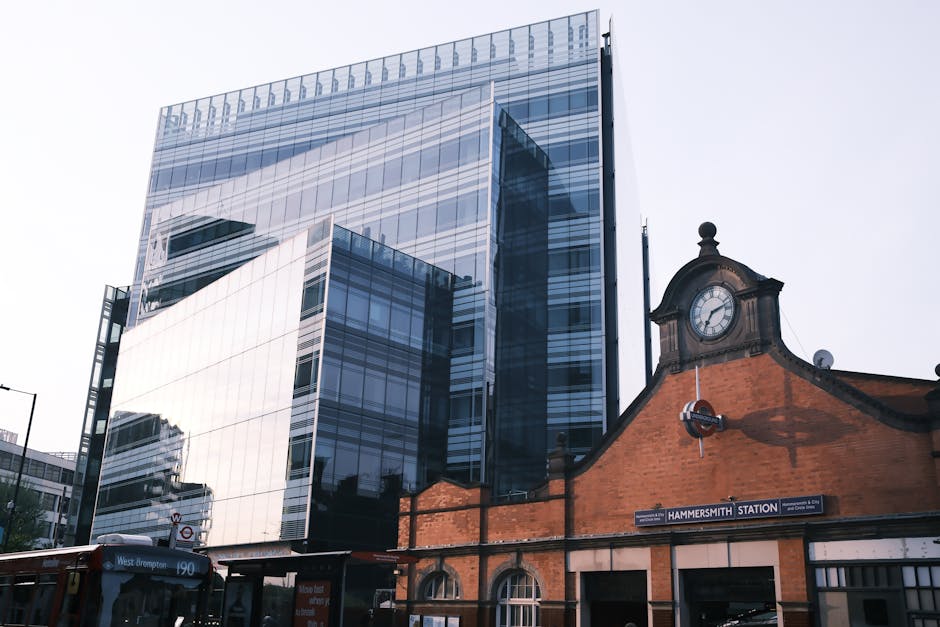 A view of a residential suburban area with red-brick houses featuring tiled roofs and chimneys in the foreground, surrounded by lush green trees. In the background, a cityscape of modern high-rise buildings and office towers extends across the horizon under a clear sky. The image is framed by overhanging tree branches at the top, and the scene appears to be taken from an elevated vantage point, possibly during daylight hours. This setting reflects an urban environment that could relate to house removals or relocation services, with visible housing and city infrastructure.