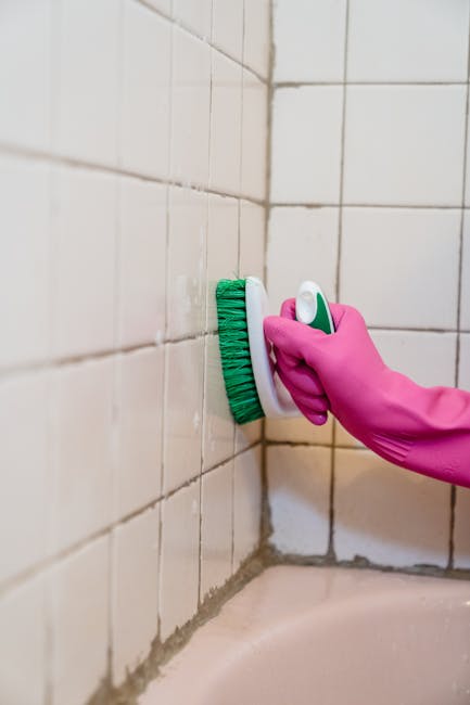 A person wearing pink rubber gloves holding a green and white scrub brush while cleaning white ceramic wall tiles in a bathroom. The tiles are arranged in a grid pattern with grout lines visible, and some areas appear to be freshly scrubbed. The background shows the tiled wall extending upward and the edge of a bathtub or sink surface at the bottom of the image. This cleaning action highlights the preparation process for home relocation, moving, or packing activities, as part of general household maintenance during a house move, as handled by Man with Van Wormwood Scrubs. The lighting is bright, emphasizing the cleanliness and care taken during the cleaning and moving preparations.