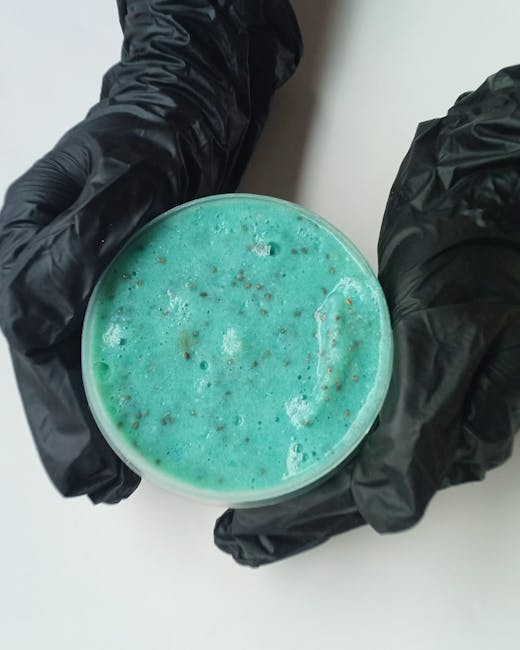 A person wearing black disposable gloves is holding a clear plastic cup filled with a blue-green liquid that has bubbles and foamy foam on the surface. The background consists of a plain white surface, and the image appears to be taken indoors, possibly during packing or cleaning tasks related to house removals. The focus is on the cup and the gloves, with no additional furniture or packaging materials visible in the image. This visual could relate to cleaning or packing liquids used during a home relocation process, as documented by Man with Van Wormwood Scrubs in their house removal services for the Scrubs Lane to White City area.