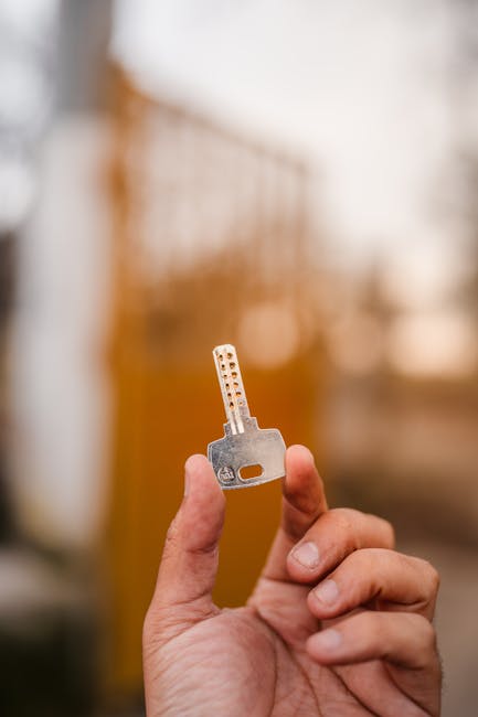 A close-up of a person's hand holding a metal key with multiple cut-outs and a rectangular hole, positioned in front of a blurred wooden staircase with vertical balusters in a residential property. Natural light illuminates the scene, highlighting the key and the hand, as part of a home relocation or moving process. This image showcases the key relevant to house removals and furniture transport services offered by Man with Van Wormwood Scrubs, emphasizing the importance of secure access during packing, loading, and personal property transfer through steps and doorways.