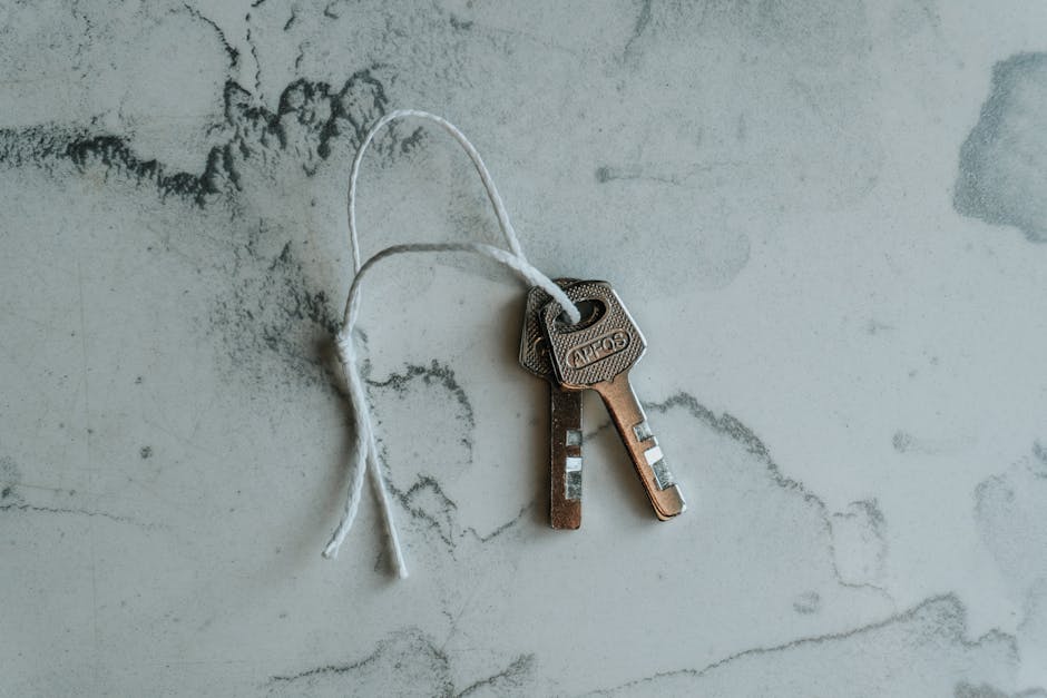 A pair of metallic house keys attached to a white string is placed on a light grey, textured surface with dark marbling and subtle cracks. The keys are standard in design, with one slightly longer than the other, and both have rectangular heads with the brand name 'COUZ' engraved on them. The keys are positioned close to each other, with the string loosely looped through their holes. The background suggests an indoor setting, possibly near an entryway, and the image captures the keys in a flat, overhead perspective. This visual supports house relocation and moving services, emphasizing the preparation and key transfer aspects involved in home removals performed by companies like Man with Van Wormwood Scrubs, facilitating efficient furniture transport and packing logistics during a property move.