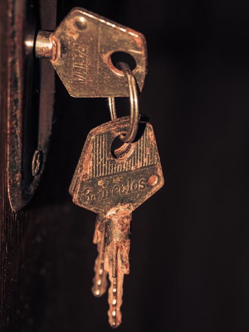 A close-up of a person's hand holding a metal key with multiple cut-outs and a rectangular hole, positioned in front of a blurred wooden staircase with vertical balusters in a residential property. Natural light illuminates the scene, highlighting the key and the hand, as part of a home relocation or moving process. This image showcases the key relevant to house removals and furniture transport services offered by Man with Van Wormwood Scrubs, emphasizing the importance of secure access during packing, loading, and personal property transfer through steps and doorways.