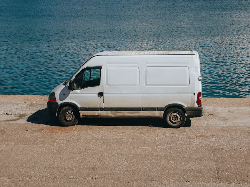 A white commercial van parked on a concrete surface beside a body of water during daylight, with the van facing to the left. The van's side panels are plain, with no visible branding or signage, and it has black bumpers and side mirrors. The scene shows the side profile of the vehicle, capturing part of the front, middle, and rear sections, with shadows cast by sunlight. The background features calm water with gentle ripples, indicating a lakeside or riverside location. The setting appears suitable for the loading or unloading of household or office items, consistent with home relocation or furniture transport activities. The image illustrates the vehicle used by Man with Van Wormwood Scrubs for professional removals and moving services, emphasizing the importance of secure vehicle parking and careful handling during packing and moving operations.