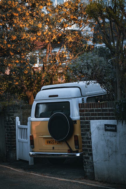 A vintage yellow and white camper van is parked partially behind a brick wall and a white gate on a residential street, with a spare tire mounted on the rear door. The van is positioned close to a white fence and a brick column, with the rear end protruding onto the narrow pavement. Surrounding the scene are tall trees with green and autumnal leaves, some of which are illuminated by warm sunlight, casting soft shadows. Visible building elements include the brick wall, a house number sign reading 'Tanglewood,' and a taller structure behind the trees. The image captures a typical home relocation or furniture transport scenario involving a vehicle prepared for moving household goods, with the van ready for loading or unloading in an outdoor environment near homes in W12, as part of house removal services offered by Man with Van Wormwood Scrubs. The environment appears calm, with natural lighting emphasizing the vehicles and surroundings.