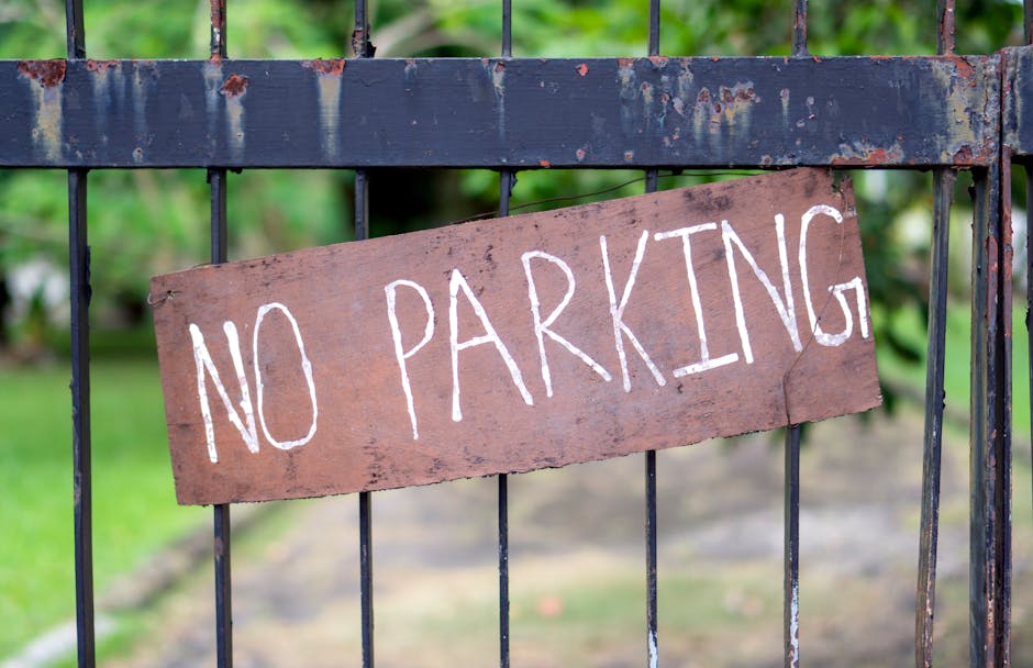 A close-up view of a black metal gate with vertical bars and a horizontal support bar, featuring a weathered, rectangular wooden sign attached with visible nails. The sign has a reddish-brown hue and displays hand-painted white capital letters reading 'NO PARKING.' The background shows a blurred garden area with green foliage and a paved path, suggesting an outdoor setting. The scene is illuminated with natural daylight, and the overall context relates to restrictions or regulations that could impact home relocation or furniture transport services provided by Man with Van Wormwood Scrubs, with the sign indicating a parking suspension or prohibition near the property.
