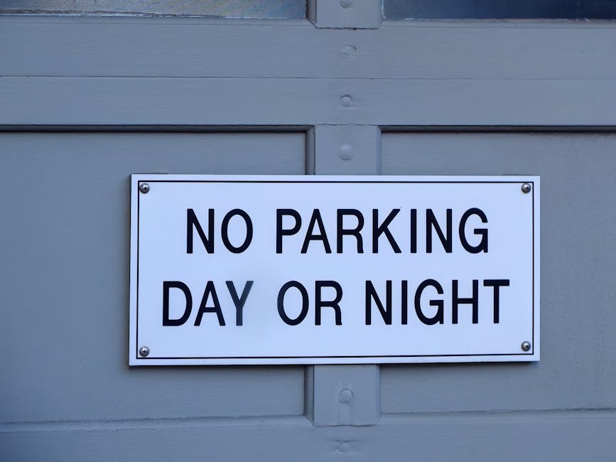A close-up view of a black metal gate with vertical bars and a horizontal support bar, featuring a weathered, rectangular wooden sign attached with visible nails. The sign has a reddish-brown hue and displays hand-painted white capital letters reading 'NO PARKING.' The background shows a blurred garden area with green foliage and a paved path, suggesting an outdoor setting. The scene is illuminated with natural daylight, and the overall context relates to restrictions or regulations that could impact home relocation or furniture transport services provided by Man with Van Wormwood Scrubs, with the sign indicating a parking suspension or prohibition near the property.