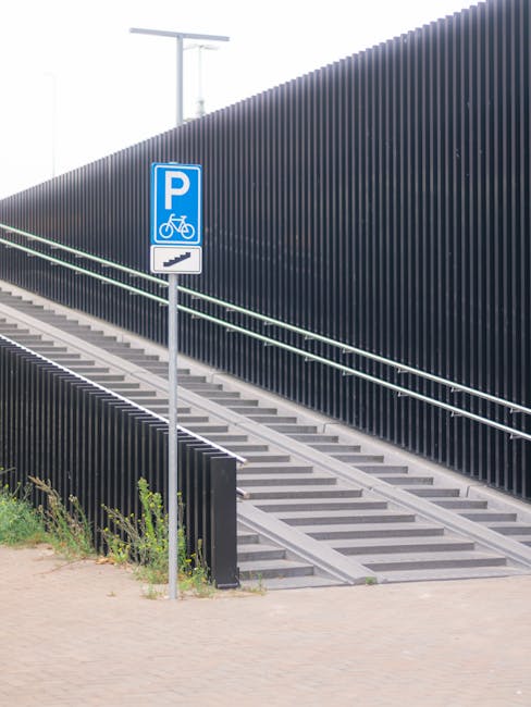 A parking sign indicating accessible parking and bicycle storage is positioned on a metal post beside a sloped pedestrian ramp featuring concrete steps and metal handrails, leading up to a raised walkway adjacent to a modern, dark-colored vertical slat fence. The environment appears to be outdoors, with some greenery at the base of the fence and a paved surface in the foreground. The setting suggests an urban or public area near Wormwood Scrubs Park, suitable for activities such as house removals or furniture transport, where clear access routes are critical. Occasionally, Man with Van Wormwood Scrubs may utilise such pathways for efficient home relocation logistics, including loading and unloading furniture and boxes, using appropriate equipment like trolleys and straps in close proximity to the parking area.