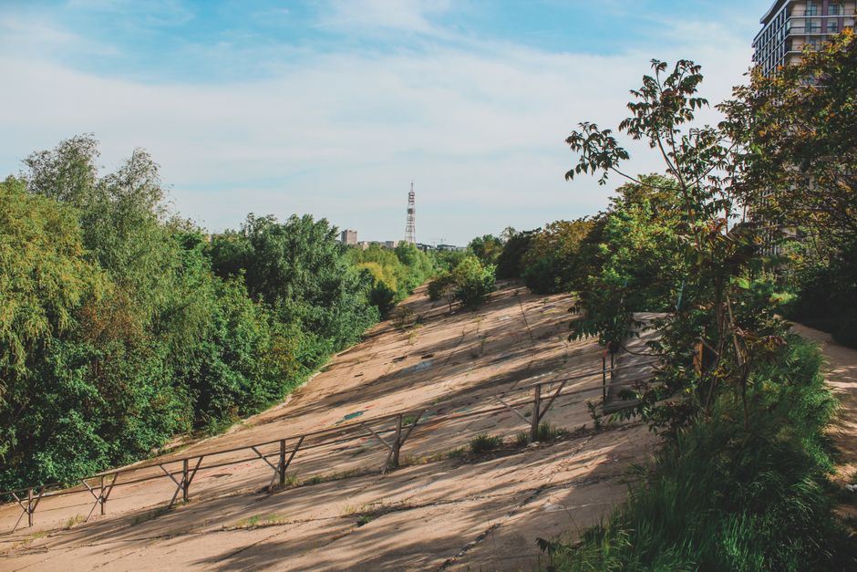A wide view of Wormwood Scrubs park showing a steep, sandy slope with a simple metal railing along the edge. The area is surrounded by dense green trees and bushes, with some taller trees on the right and more foliage in the background. On the left, the park transitions into a more open space with a clear sky overhead and soft sunlight illuminating the scene. In the distance, a communication tower is visible, along with several distant high-rise buildings on the horizon. This outdoor environment depicts a natural setting with minimal human activity, suitable for illustrating a location where a home relocation or furniture transport process might take place near a public park. The scene emphasizes the natural landscape with the sandy slope as a key feature, providing context for logistics and moving services in the Wormwood Scrubs area, as offered by Man with Van Wormwood Scrubs.