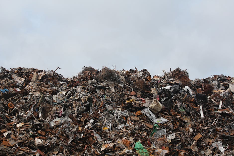 A large pile of mixed waste materials, including broken wood, plastic, metal, cardboard, and fabric, stacked outdoors against a cloudy sky. The debris appears to be from demolition or waste disposal and is situated near a property, possibly on a pavement or designated waste area, with no visible vehicles or people in the scene. The image depicts the type of waste that may be subject to regulations and fines at Wormwood Scrubs, as detailed on the website of Man with Van Wormwood Scrubs, a house removals and relocation service provider involved in home relocation and furniture transport.